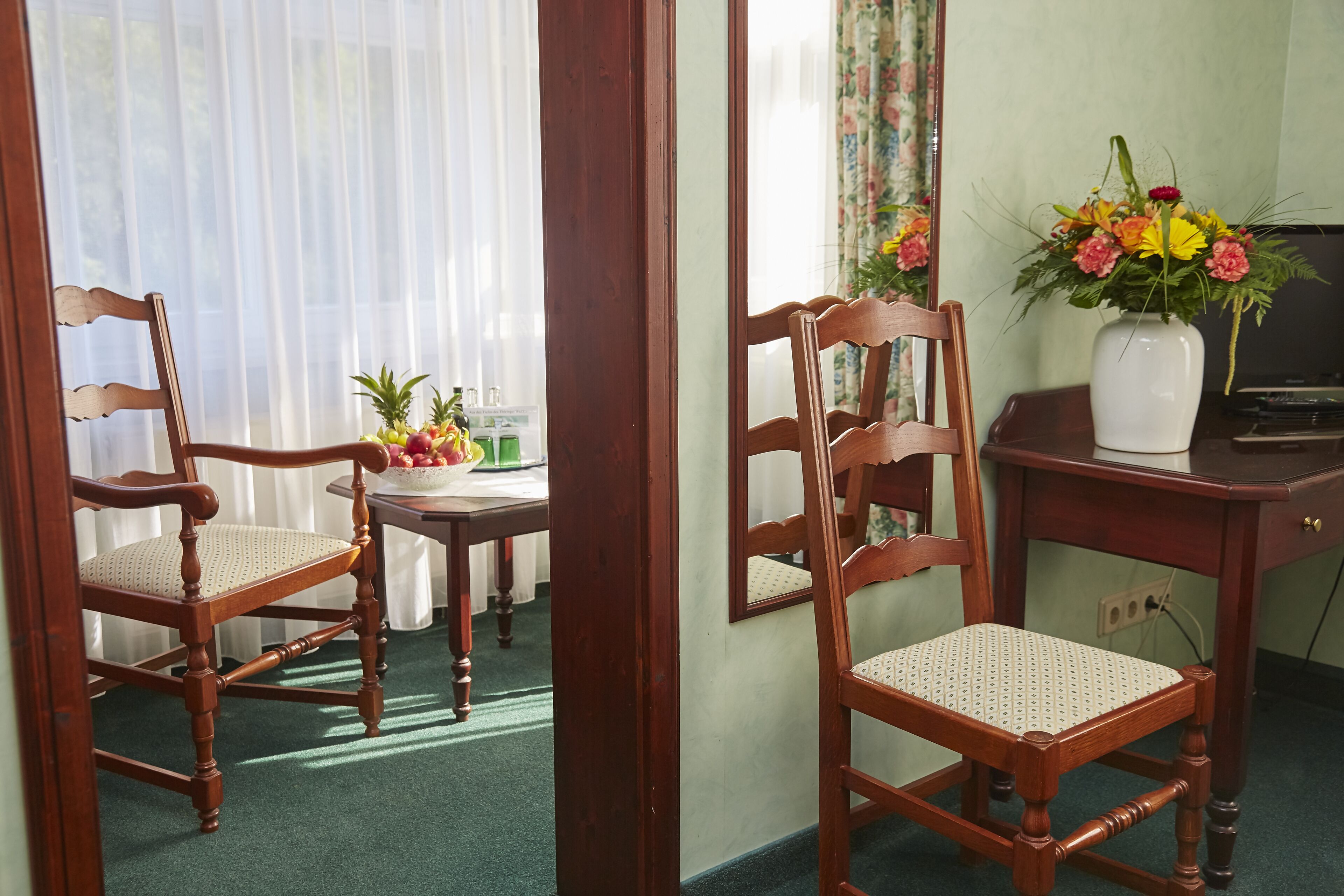 Ein Zimmer mit zwei Holzsesseln, einem kleinen Tisch mit Obstschale, einer Blumenvase und einem Spiegel an der Wand.