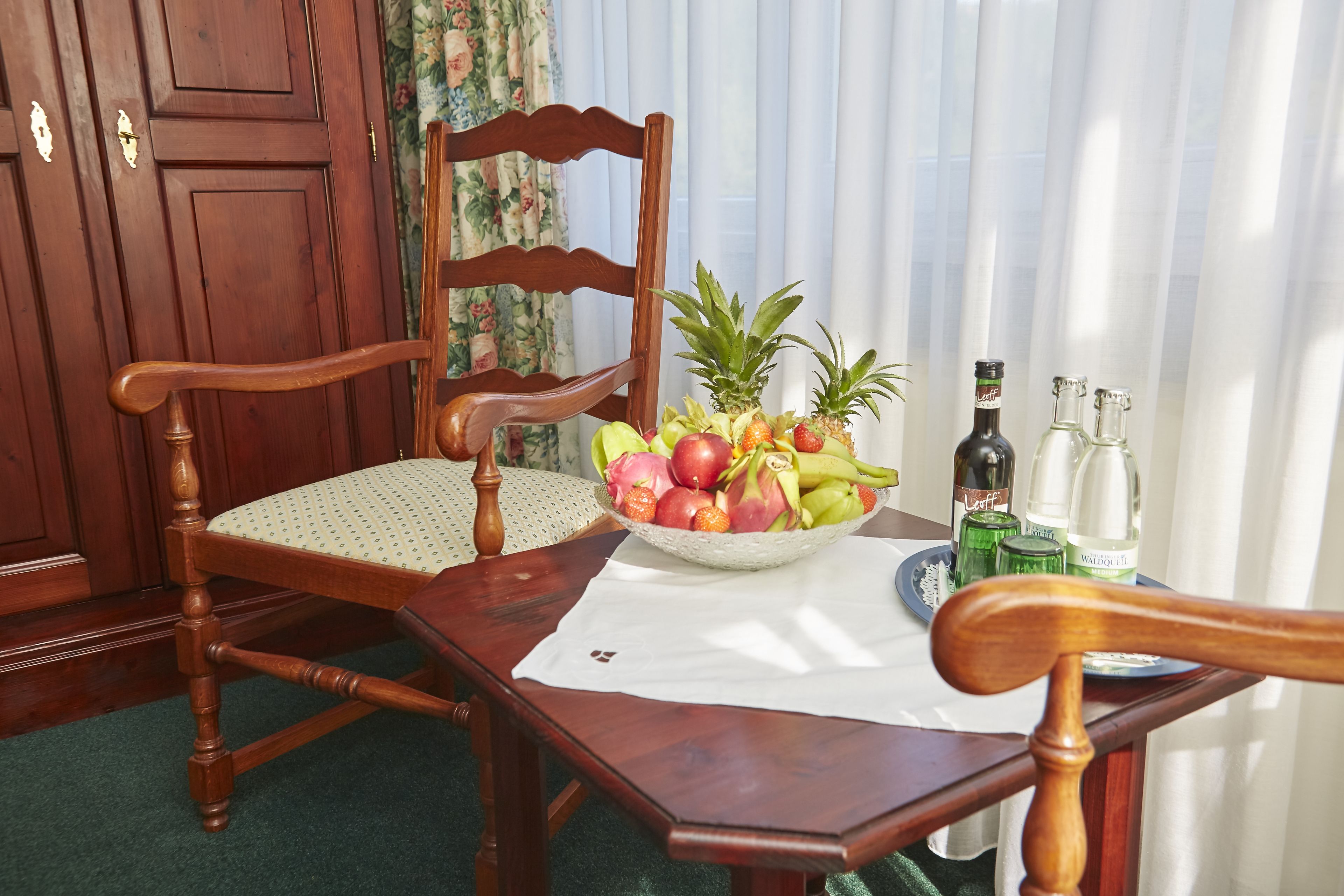 A wooden dining table with a white tablecloth, a wooden chair, a fruit bowl, and two bottles on the table.