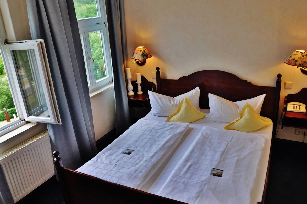 Un lit bien fait, avec du linge de lit blanc et deux oreillers, une tête de lit en bois, des lampes de chevet, une table de chevet avec un téléphone et une fenêtre avec des rideaux.