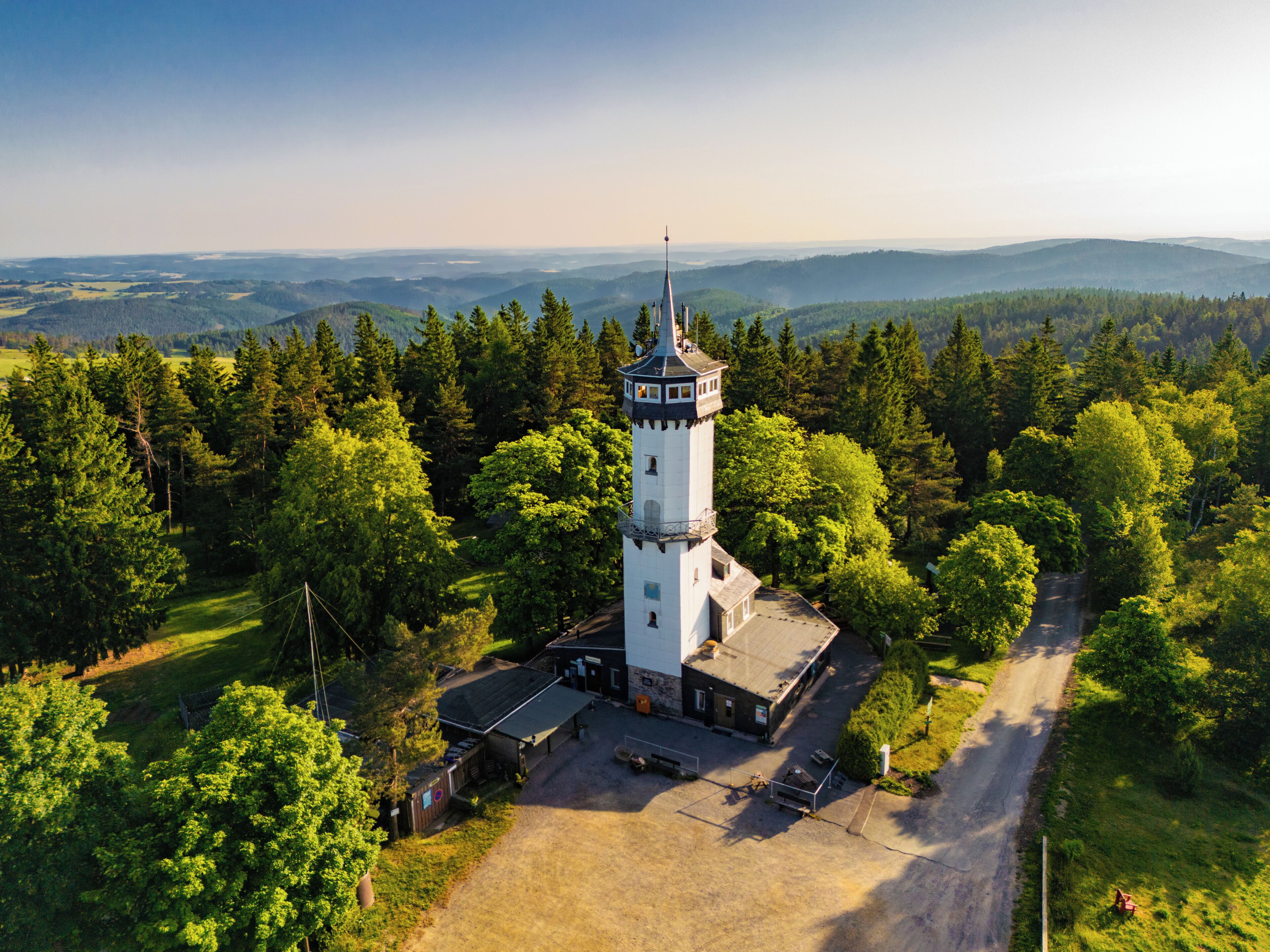 Valkoinen torni, jonka päällä on kartiomainen katto, ja sen ympärillä on tiheää metsää sekä kirkas taivas.