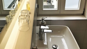 A bathroom with a sink, a mirror, and a window with a view of a house and garden. - Flair Hotel Waldfrieden (Schwarzatal)