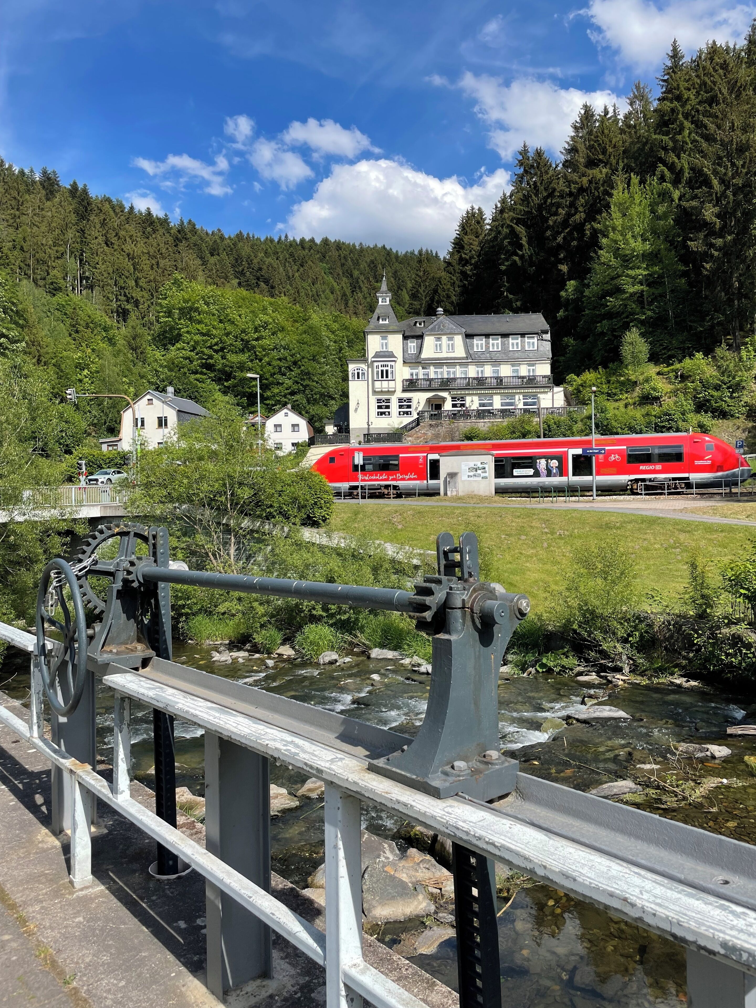 Ein roter Zug fährt an einer Brücke vorbei, unter der ein Wasserlauf fließt. Im Hintergrund ist ein großes Gebäude mit einem Balkon und ein bewaldeter Hügel zu sehen.