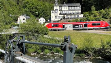 Ein roter Zug fährt an einer Brücke vorbei, unter der ein Wasserlauf fließt. Im Hintergrund ist ein großes Gebäude mit einem Balkon und ein bewaldeter Hügel zu sehen.