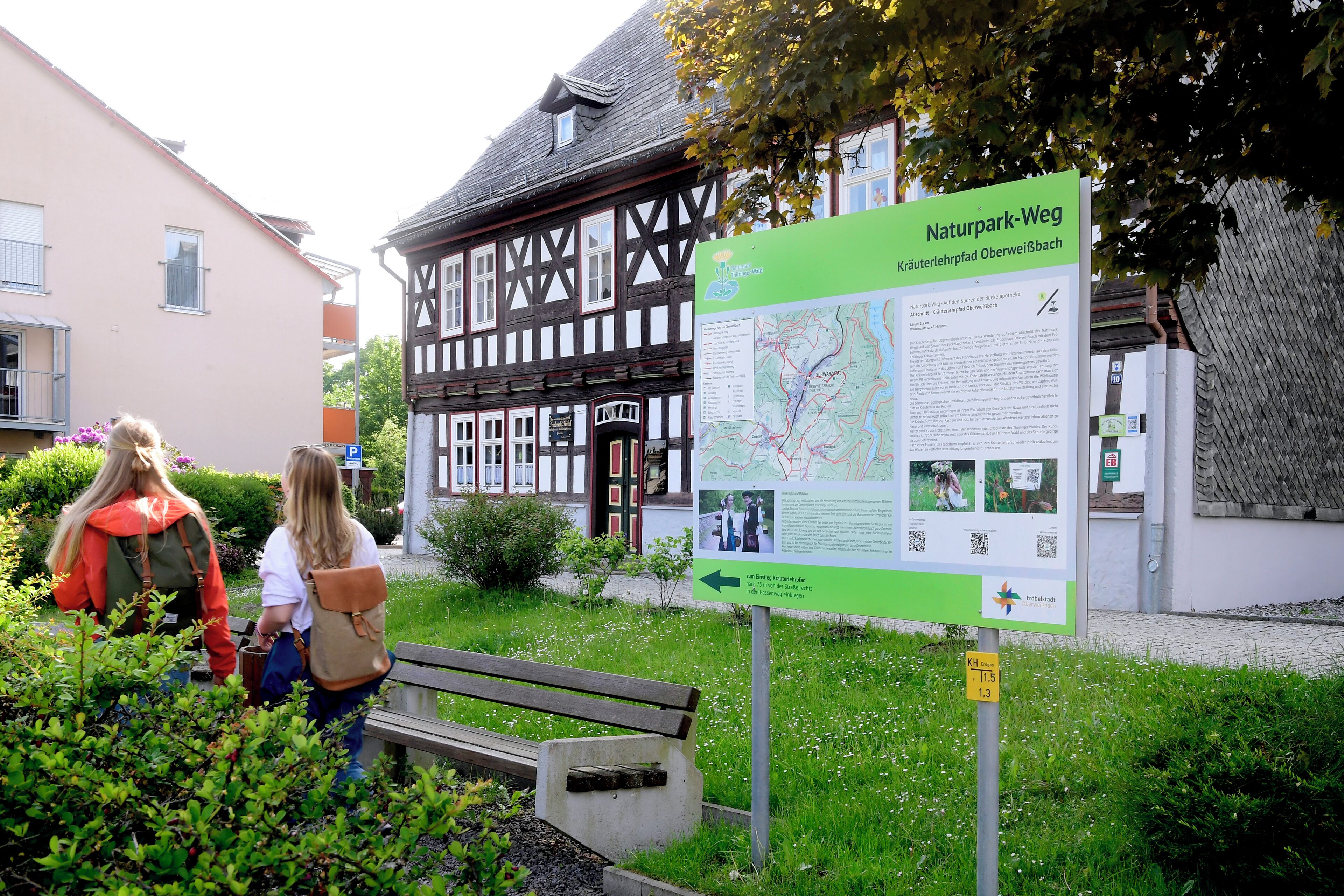 Ein historisches Gebäude mit einer traditionellen Fachwerkausfachung, einem grünen Informationsschild und einer Bank in einer angelegten Grünfläche.