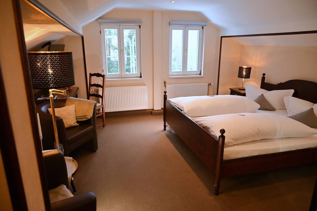 Une chambre avec un grand lit, deux fenêtres, un radiateur, une chaise et une lampe.