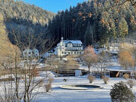 Um edifício grande, com vários andares e fachada simétrica, rodeado por terreno e árvores cobertas de neve.
