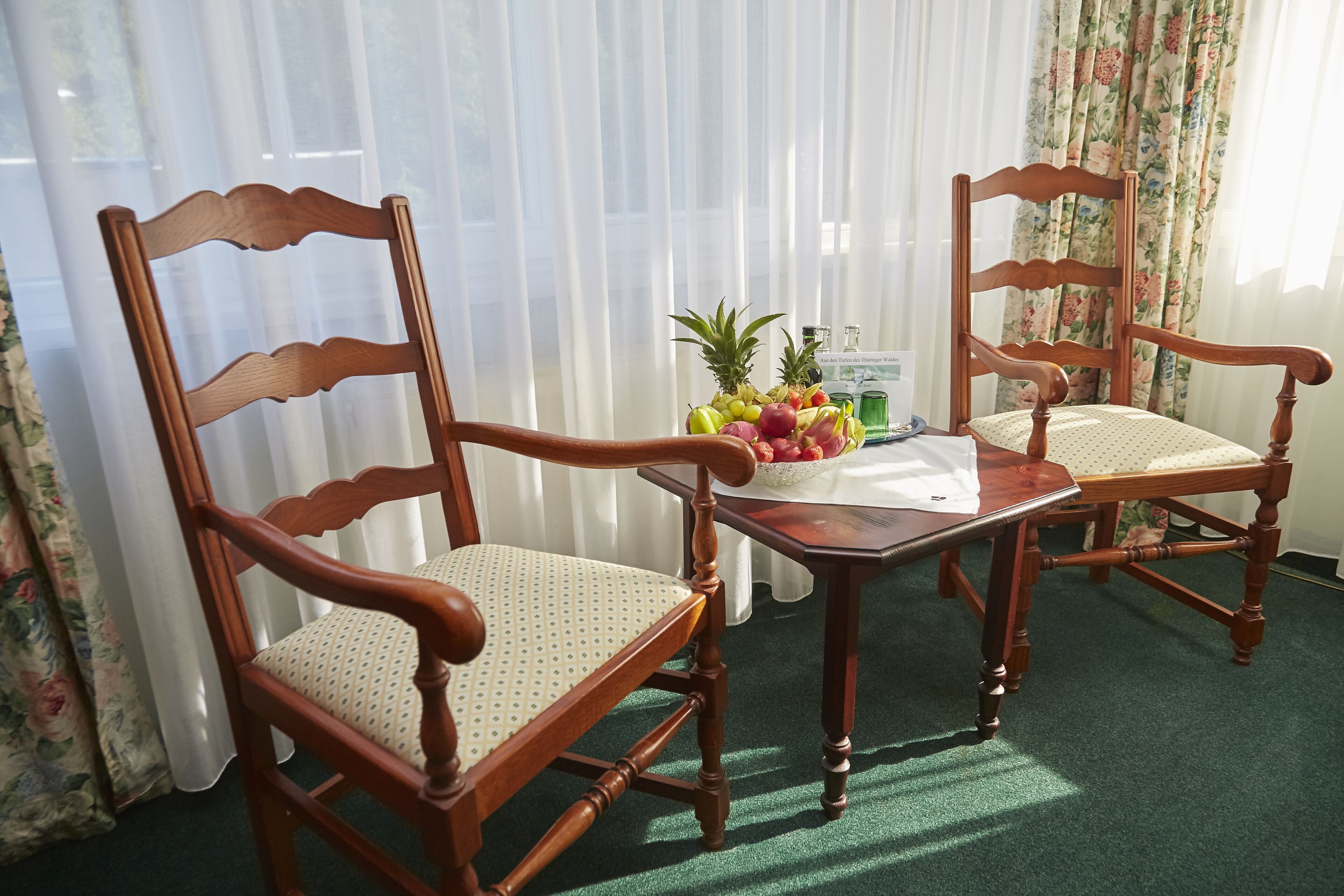 Two wooden chairs with cushioned seats and backs, a small table with a fruit bowl, and a window with sheer curtains.