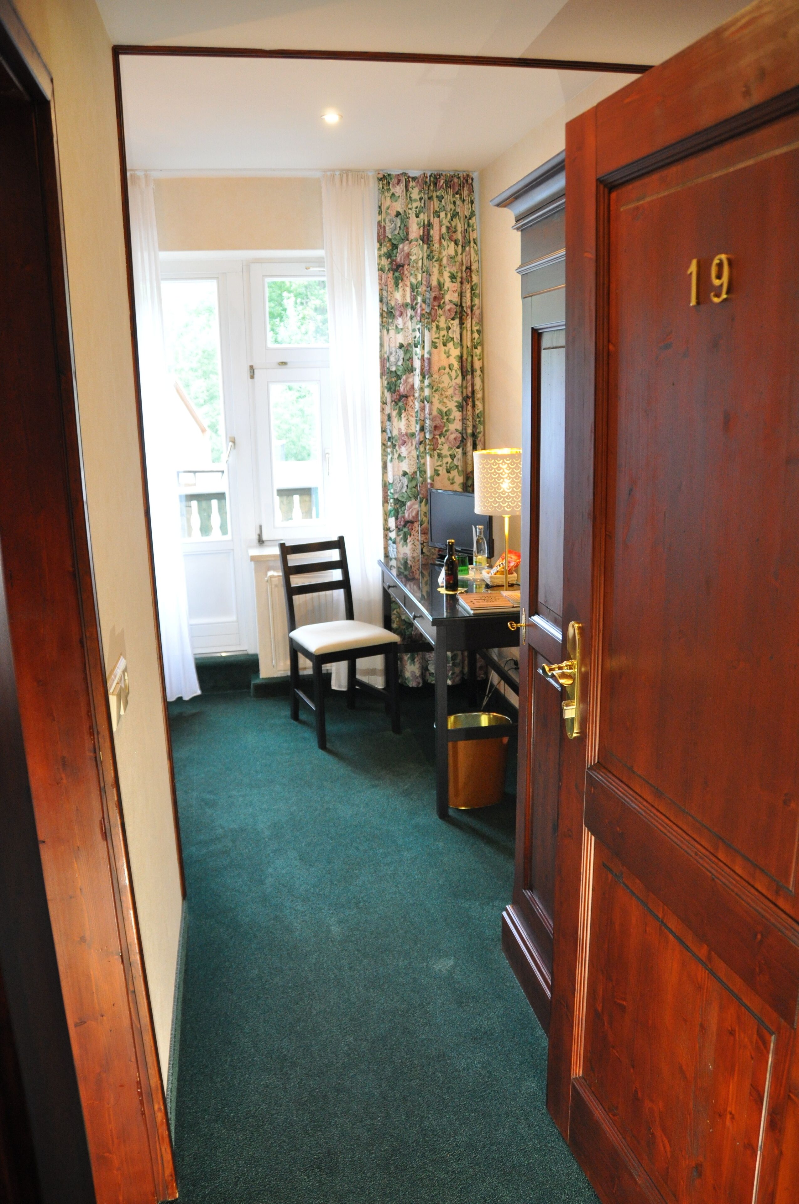 Una habitación con puerta de madera con el número 19, un escritorio con lámpara, una silla y una ventana con cortinas.
