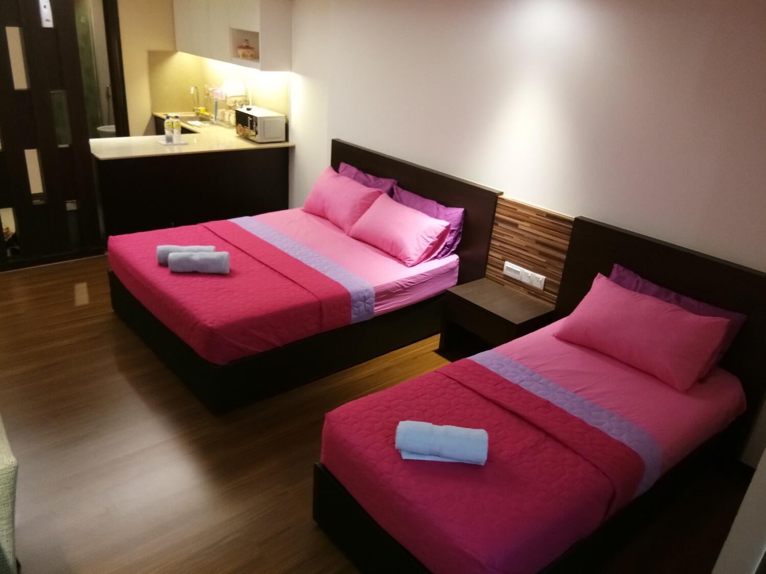 A hotel room with two beds, each with pink bedding and white towels. There is a small bedside table with a lamp and a power outlet.