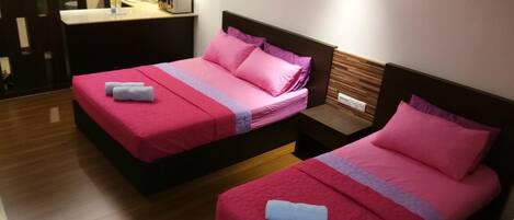 Habitación de hotel con dos camas, cada una con ropa de cama rosa y toallas blancas. Hay una mesita de noche pequeña con una lámpara y un enchufe.