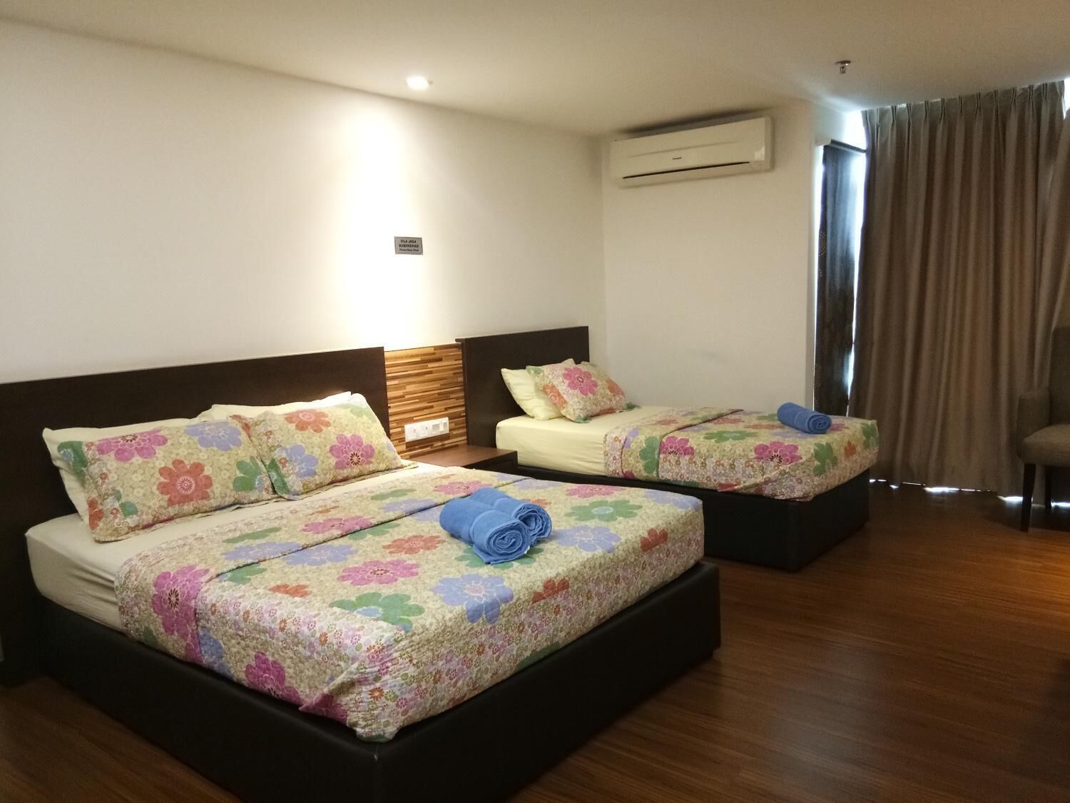 Ein Hotelzimmer mit zwei Betten, beide mit Blumenmuster bezogen und mit einem blauen Handtuch ausgestattet. Es gibt ein Holz-Kopfteil, eine Klimaanlage und ein Fenster mit Vorhängen.