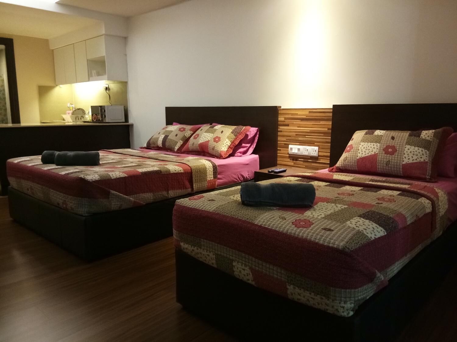 A hotel room with two beds, each with a quilted bedspread and pillows. There is a wooden headboard, a bedside table with a remote control, and a wall-mounted light.