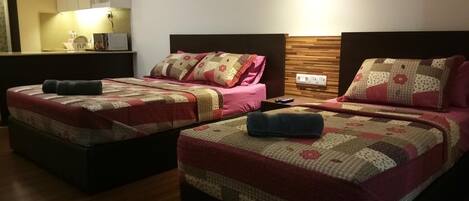 Habitación de hotel con dos camas, cada una con cubrecama acolchado y almohadas. Se observa un cabecero de madera, una mesita de noche con un control remoto y una lámpara fijada a la pared.