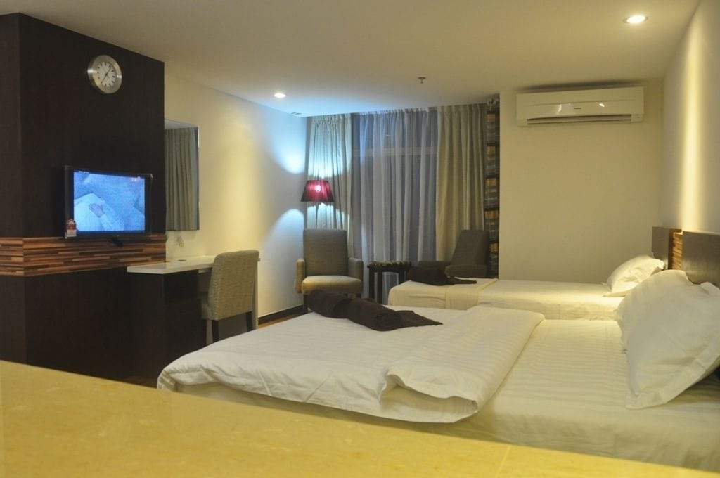 A hotel room with a bed, a desk with a television, a chair, and an air conditioning unit.