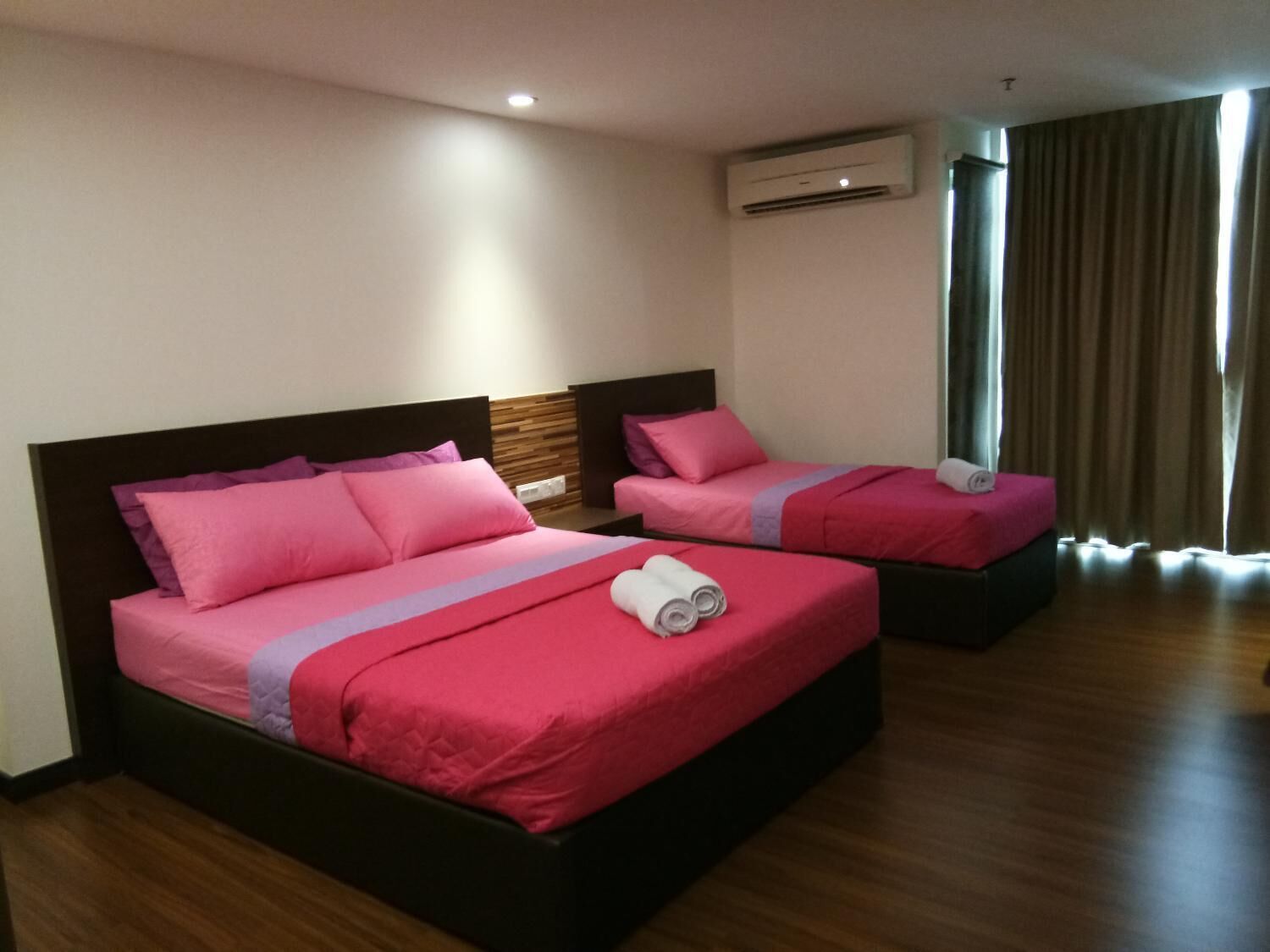 Habitación de hotel con dos camas, cada una con ropa de cama rosa y cabeceros de madera. Se observa una unidad de aire acondicionado en la pared y una ventana con cortinas.