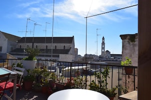 Eine Dachterrasse mit Topfpflanzen, einem Tisch und Stühlen. Im Hintergrund ein Gebäude mit einem Glockenturm.