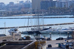 Un puerto deportivo con varios yates amarrados, una zona de estacionamiento con automóviles y un paisaje urbano al fondo.