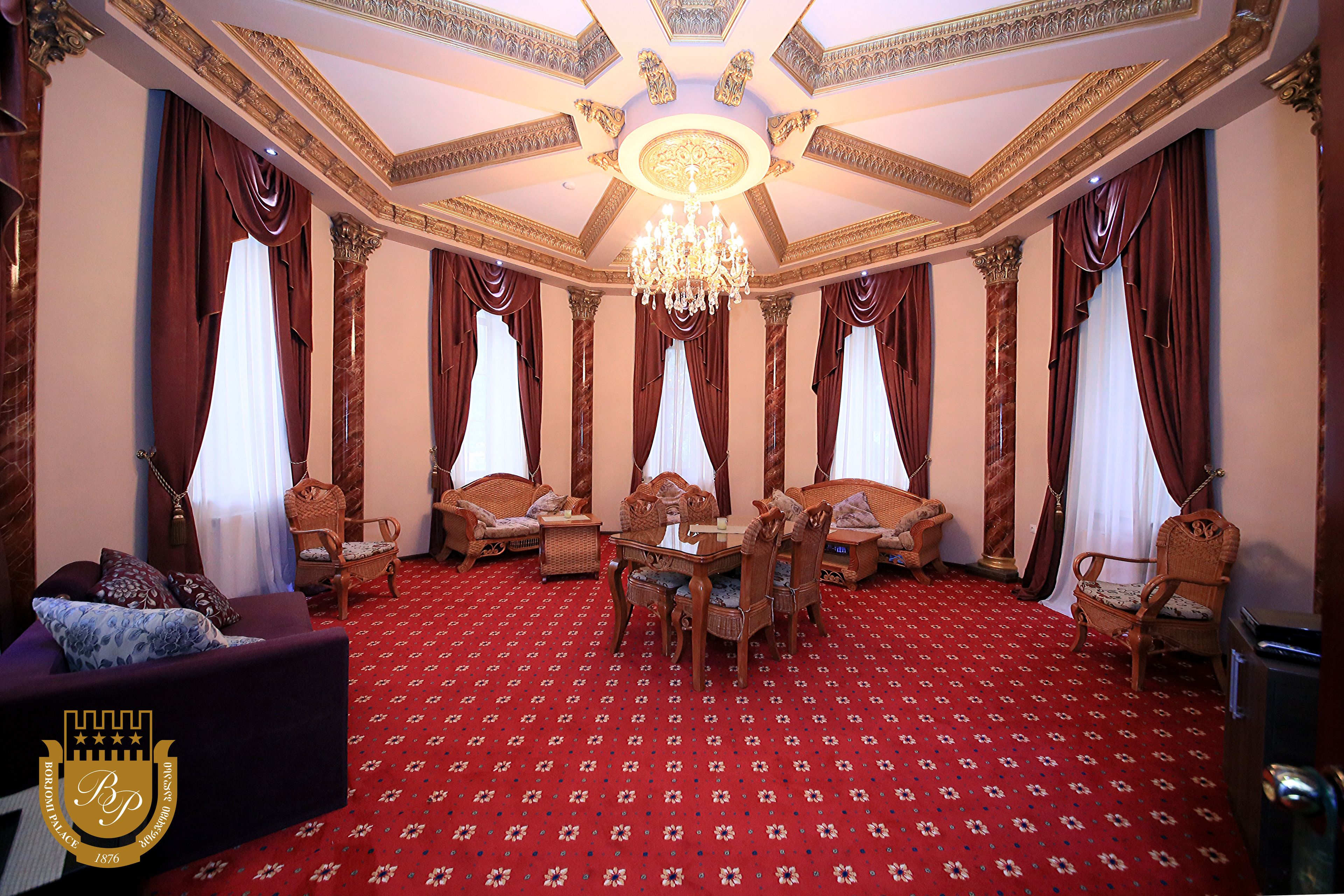 A grand room with a red carpet, ornate ceiling, and a chandelier. There are multiple seating areas with sofas and chairs.