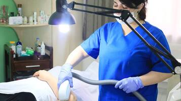 A medical professional in blue scrubs performing a procedure on a patient in a clinical setting with medical supplies and equipment visible.