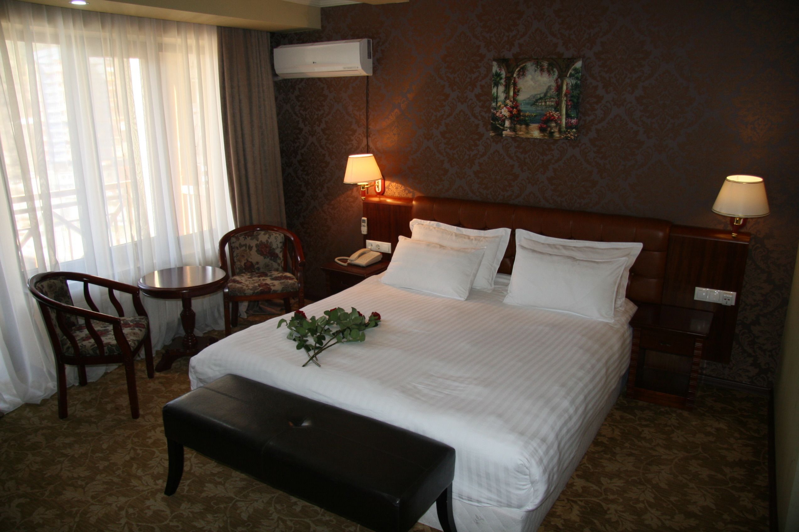 A hotel room with a large bed, two bedside lamps, a desk with a chair, and a window with sheer curtains.