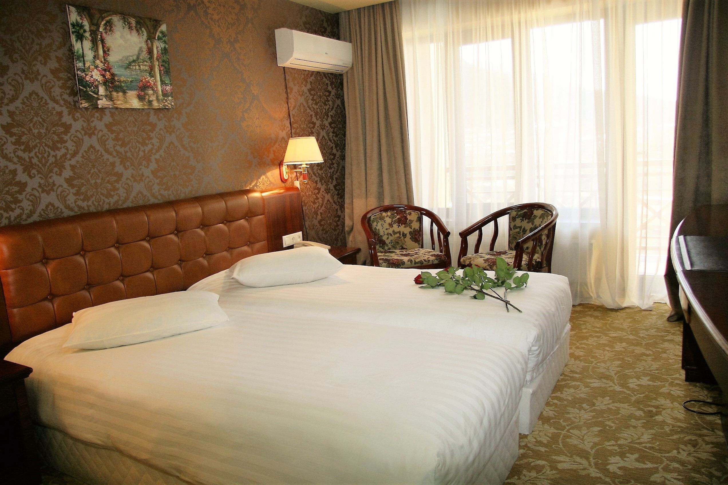 A hotel room with a large bed, a headboard, a wall-mounted air conditioner, a lamp, two chairs, and a television.