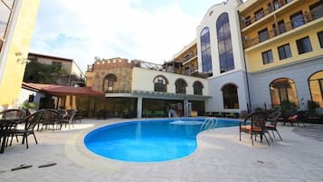 Piscina com espreguiçadeiras e mesas, cercada por edifícios com varandas e janelas em arco.