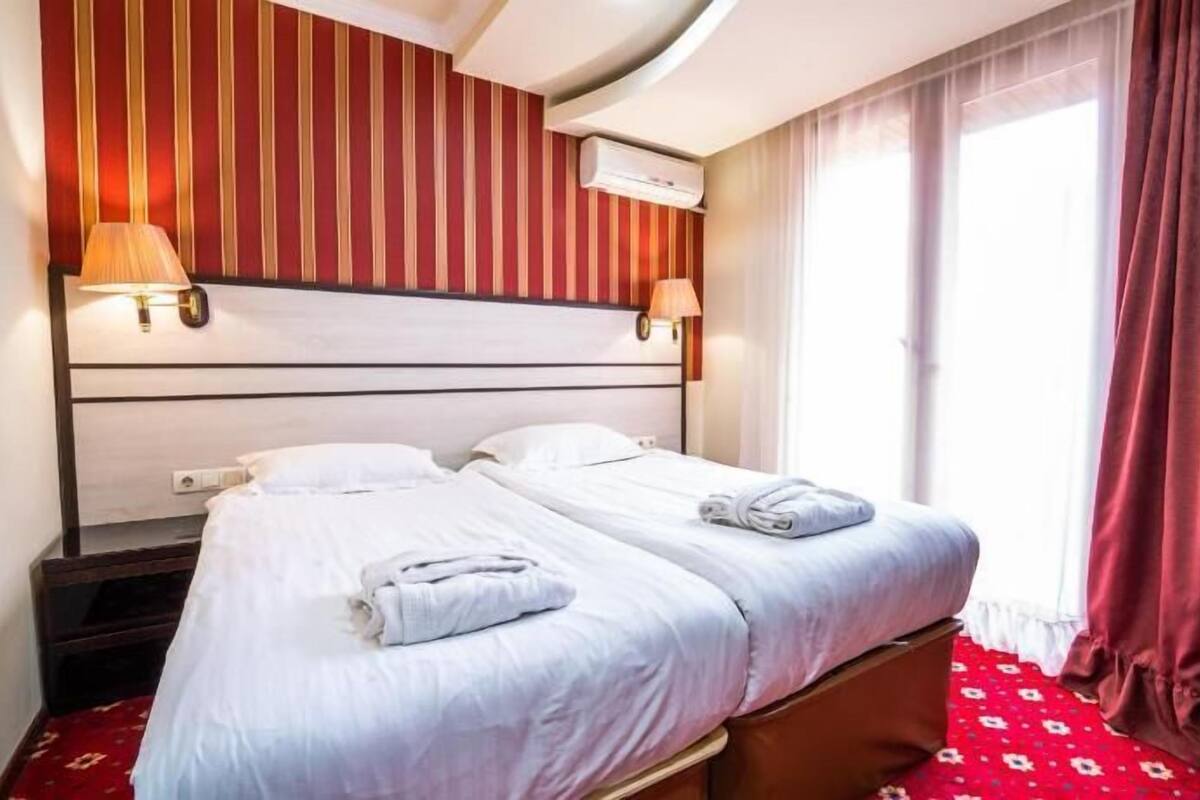 A hotel room with a bed, two bedside lamps, an air conditioning unit, and a window with curtains.