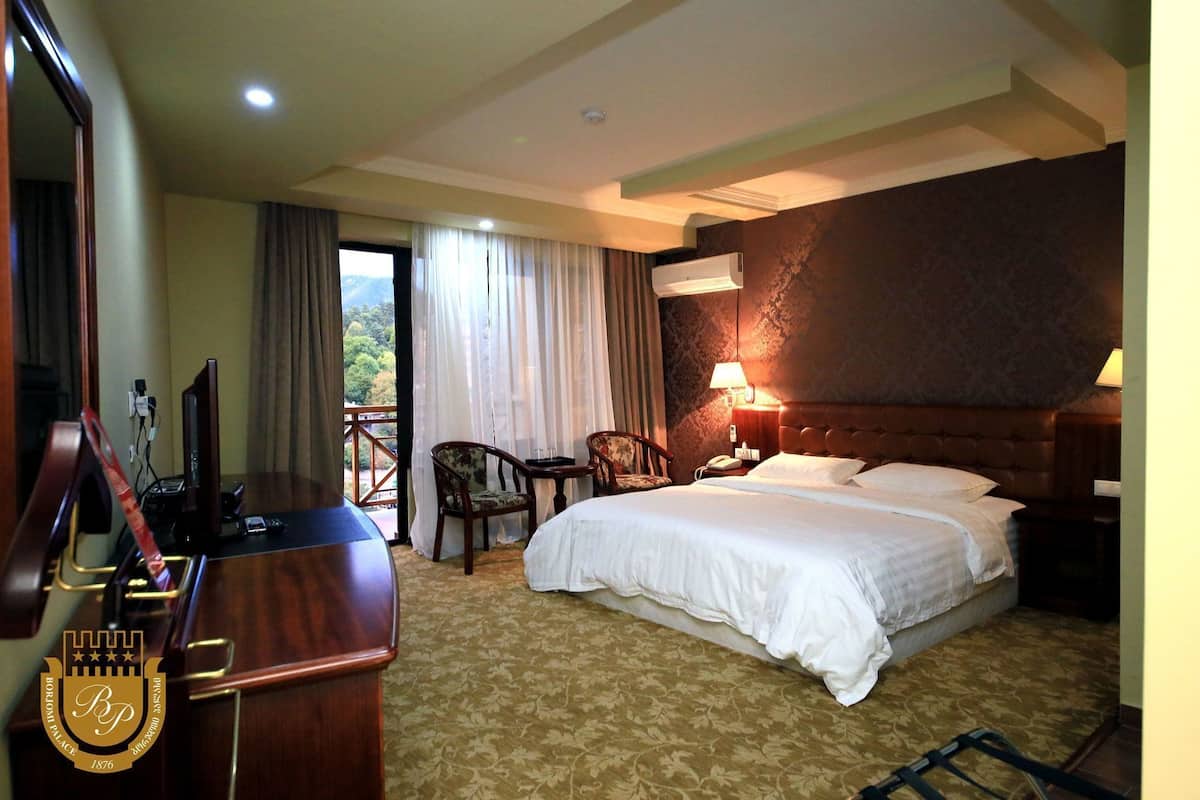 A hotel room with a bed, a desk with a computer, a chair, a nightstand, and a window with curtains.