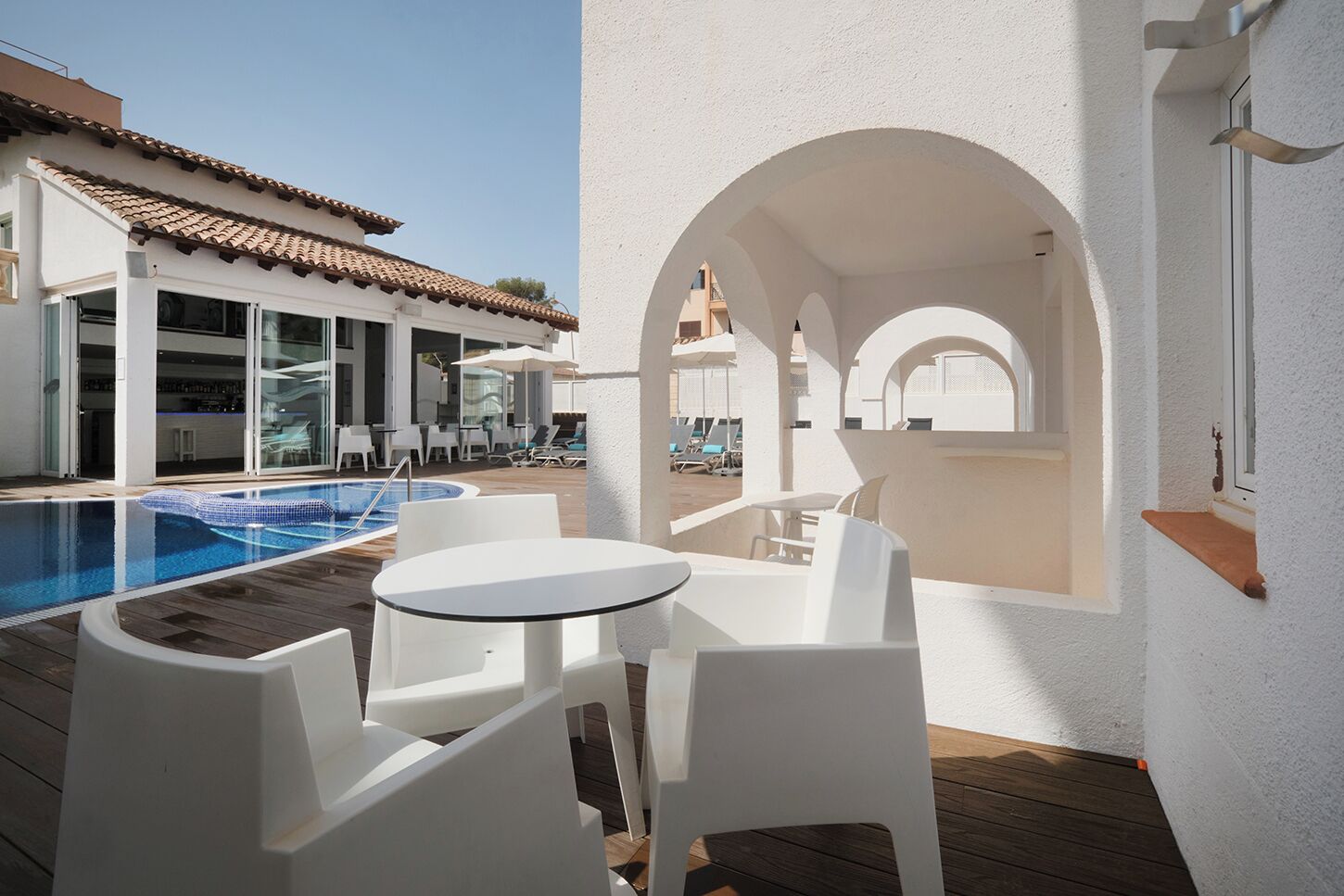 A poolside area with a pool, white chairs, and tables, and a building with a tiled roof in the background.