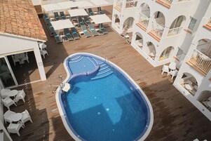 Una piscina en la azotea con tumbonas y sombrillas, rodeada de edificios blancos con balcones.