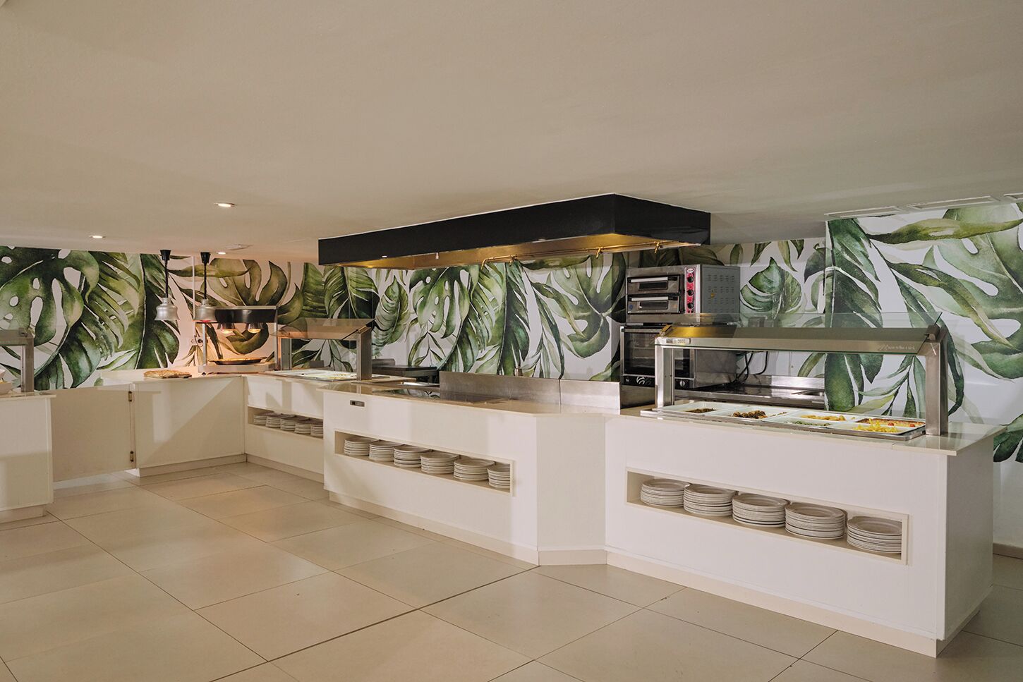 A buffet area with a tropical leaf wallpaper, a flat-screen TV, and a self-service food station with trays.