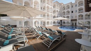Zona de piscina con tumbonas y sombrillas, piscina y un edificio de varios pisos con balcones.