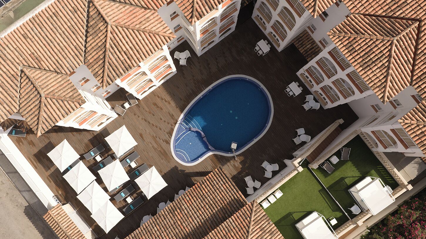 Vista aérea de un hotel con piscina rodeada de tumbonas y sombrillas, varios balcones y una zona de césped.