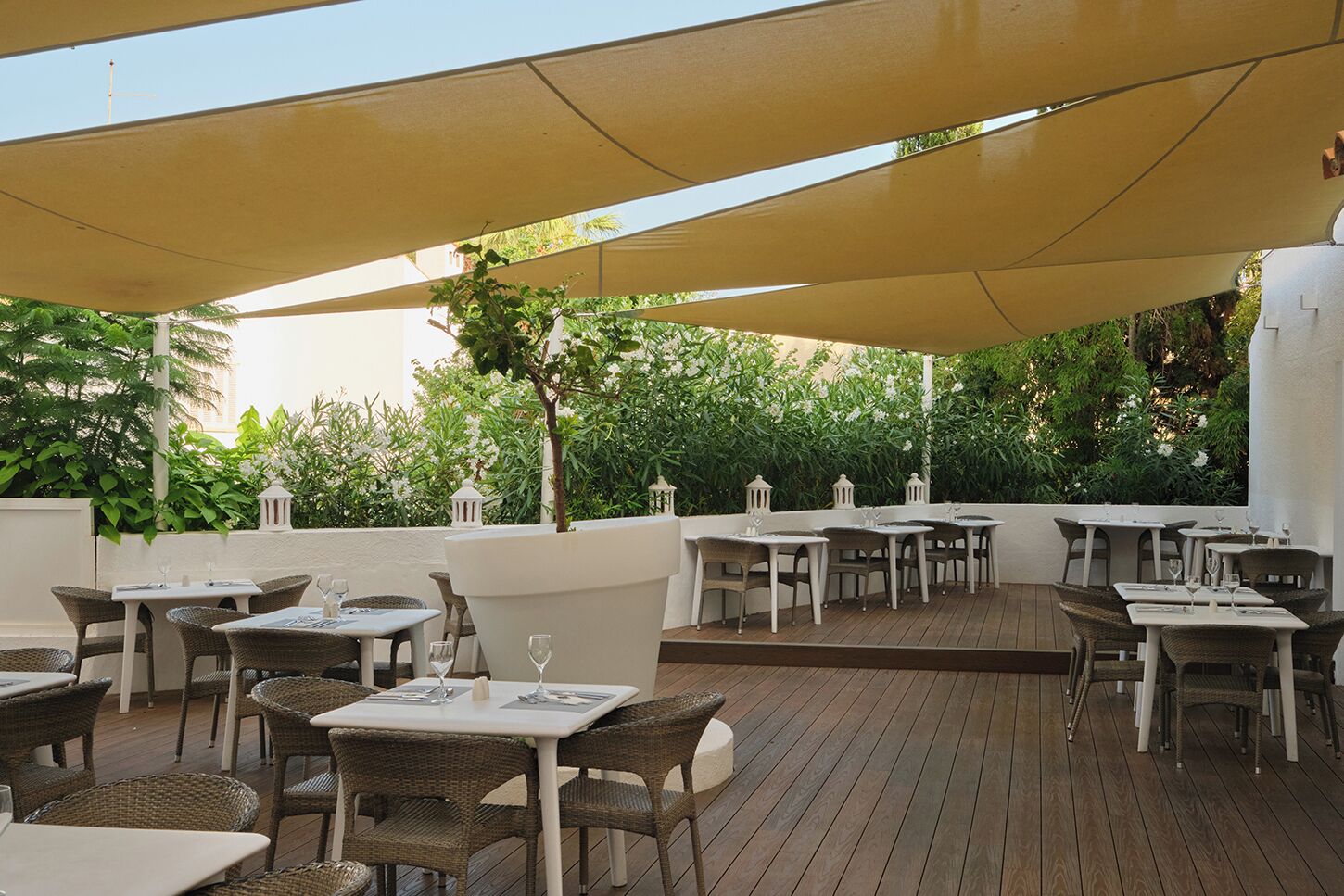 Un patio al aire libre con mesas y sillas, rodeado de vegetación y un muro blanco.