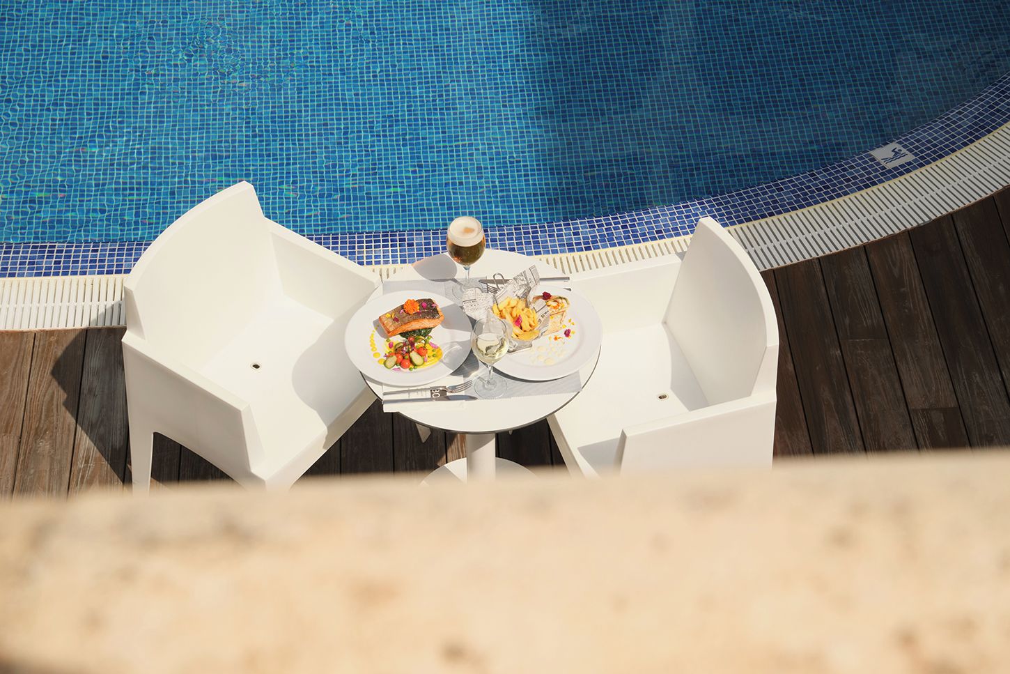 A dining table with two plates of food and a glass of wine, positioned near a pool with a blue tiled edge.