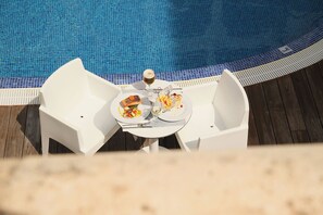 Una mesa de comedor con dos platos de comida y una copa de vino, ubicada cerca de una piscina con borde de azulejos azules.