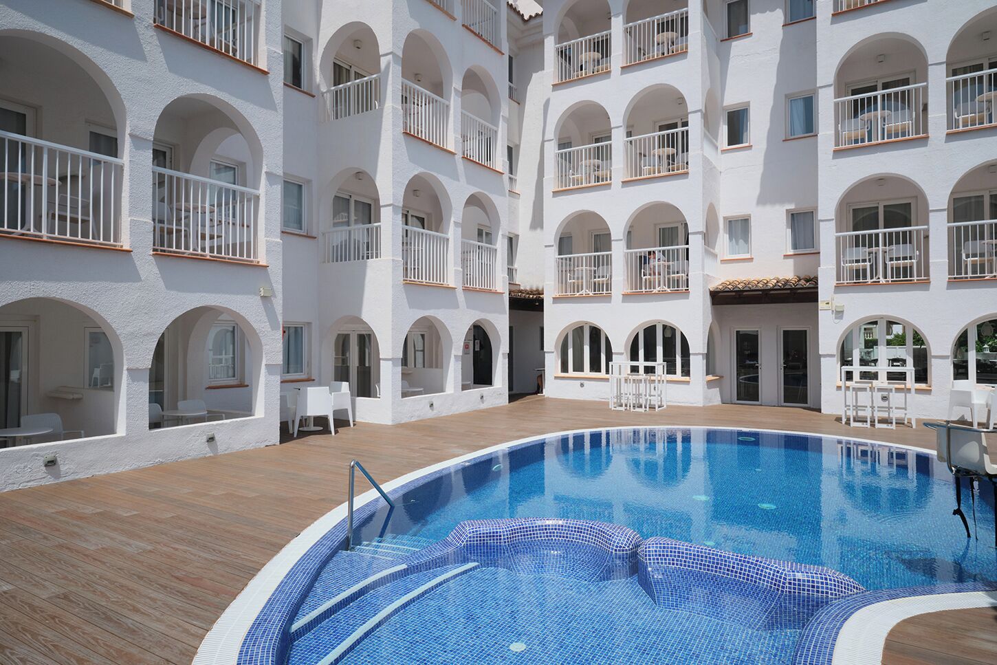 A hotel with a central swimming pool, multiple balconies with white railings, and a courtyard with outdoor seating.