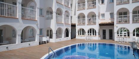 A hotel with a central swimming pool, multiple balconies with white railings, and a courtyard with outdoor seating.