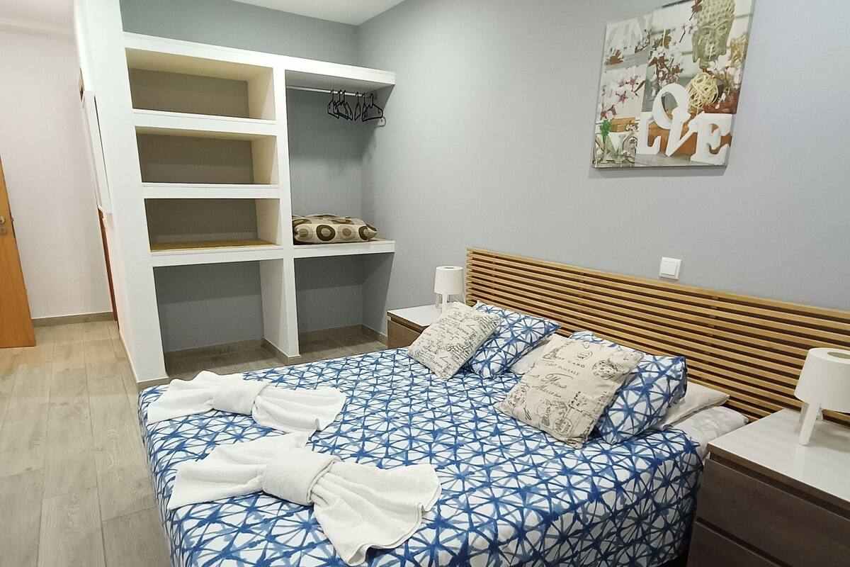 A bedroom with a bed, a wooden headboard, a nightstand, a bedside table, a wall-mounted shelf unit, and a painting.