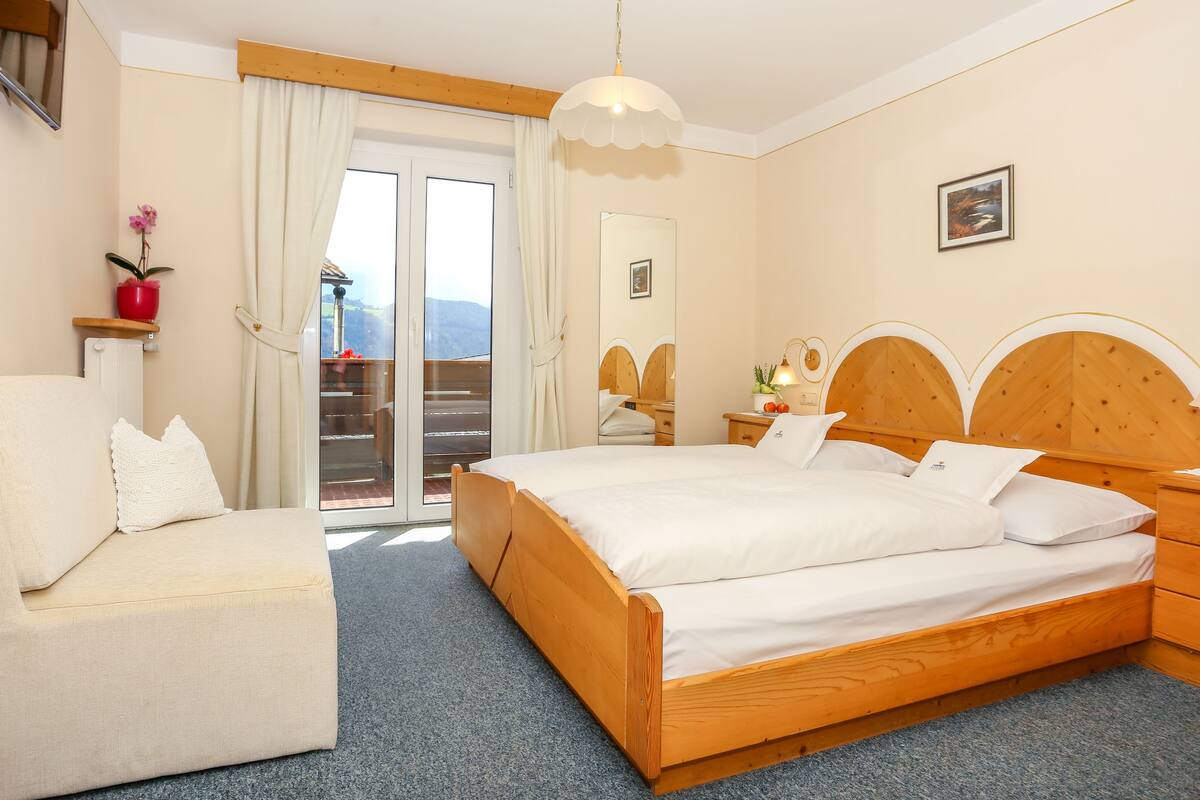 A bedroom with a wooden bed, a sofa, a small table, and a window with a view of mountains.