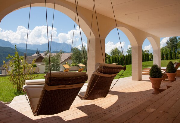A wooden deck with two wicker chairs and a swing, overlooking a green landscape and mountains. - Sonnenhotel Adler (Villandro)