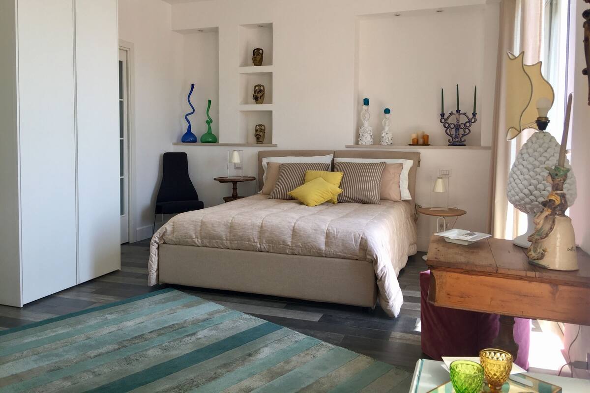 Ein Schlafzimmer mit einem Bett, Nachttischen, einem Sessel, einer Kommode und dekorativen Gegenständen auf Regalen.