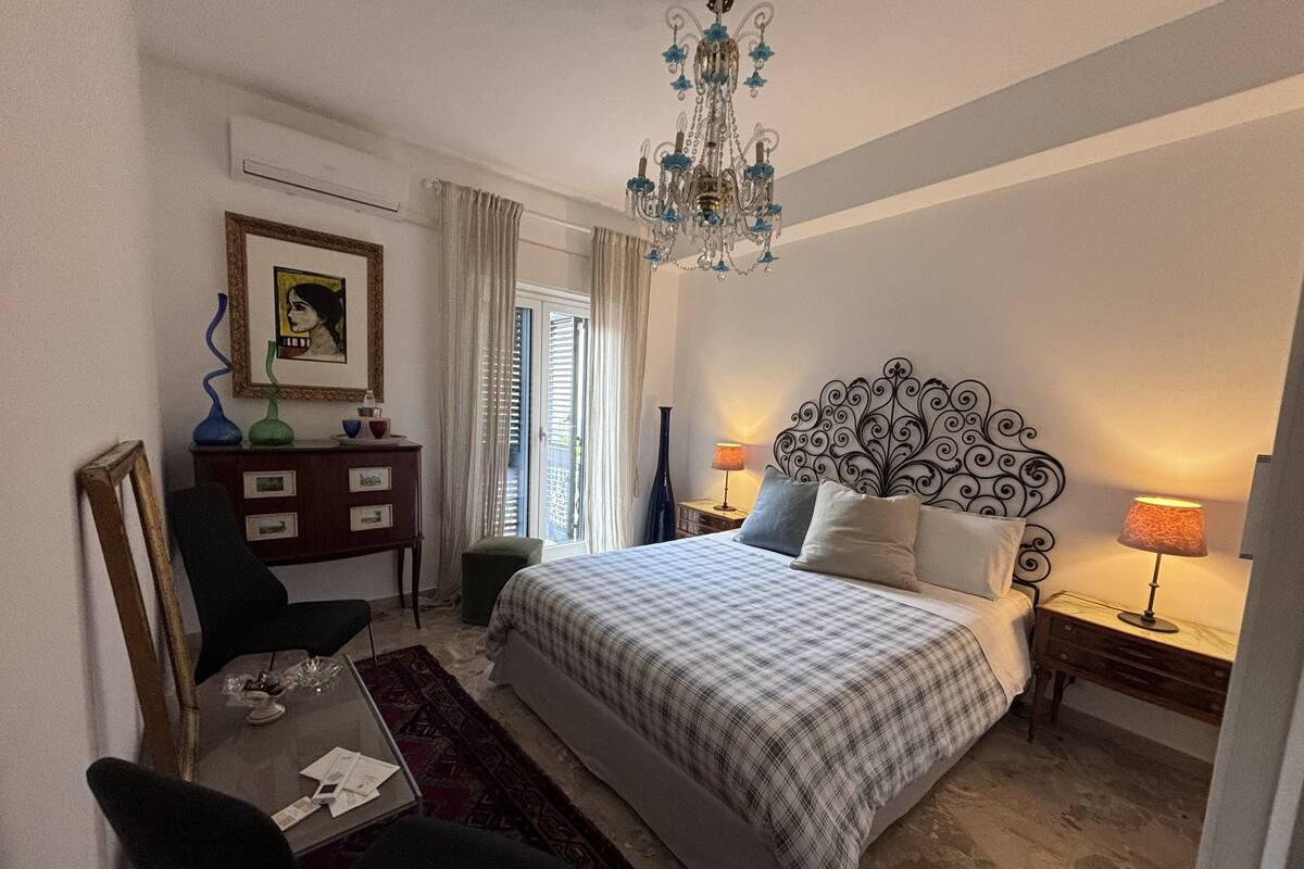 Ein Schlafzimmer mit Bett, Schreibtisch, Stuhl, Kommode, einer Kandelaber und einem Fenster mit Vorhängen.
