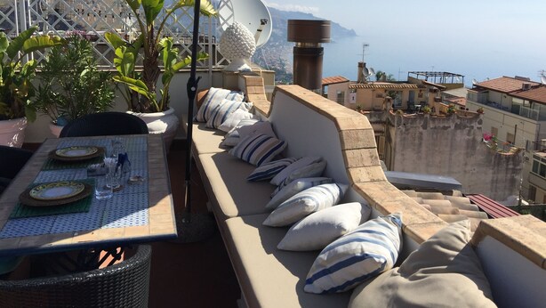 A rooftop terrace with a table set for dining, a cushioned seating area, and a large umbrella providing shade. - Casa Aricò & Shatulle Suites (Taormina)
