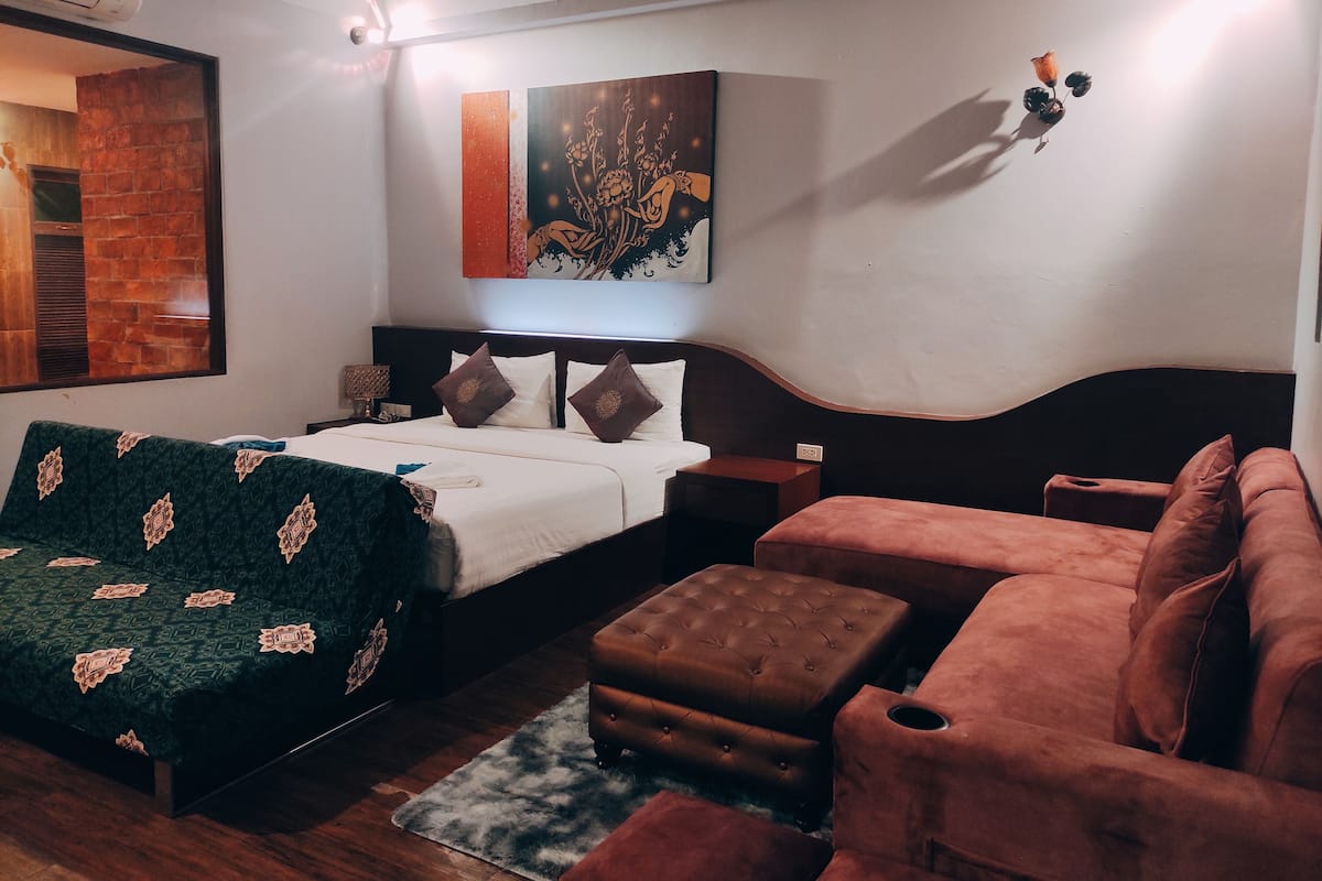 A bedroom with a bed, a sofa, an ottoman, a wall-mounted artwork, and a brick wall.