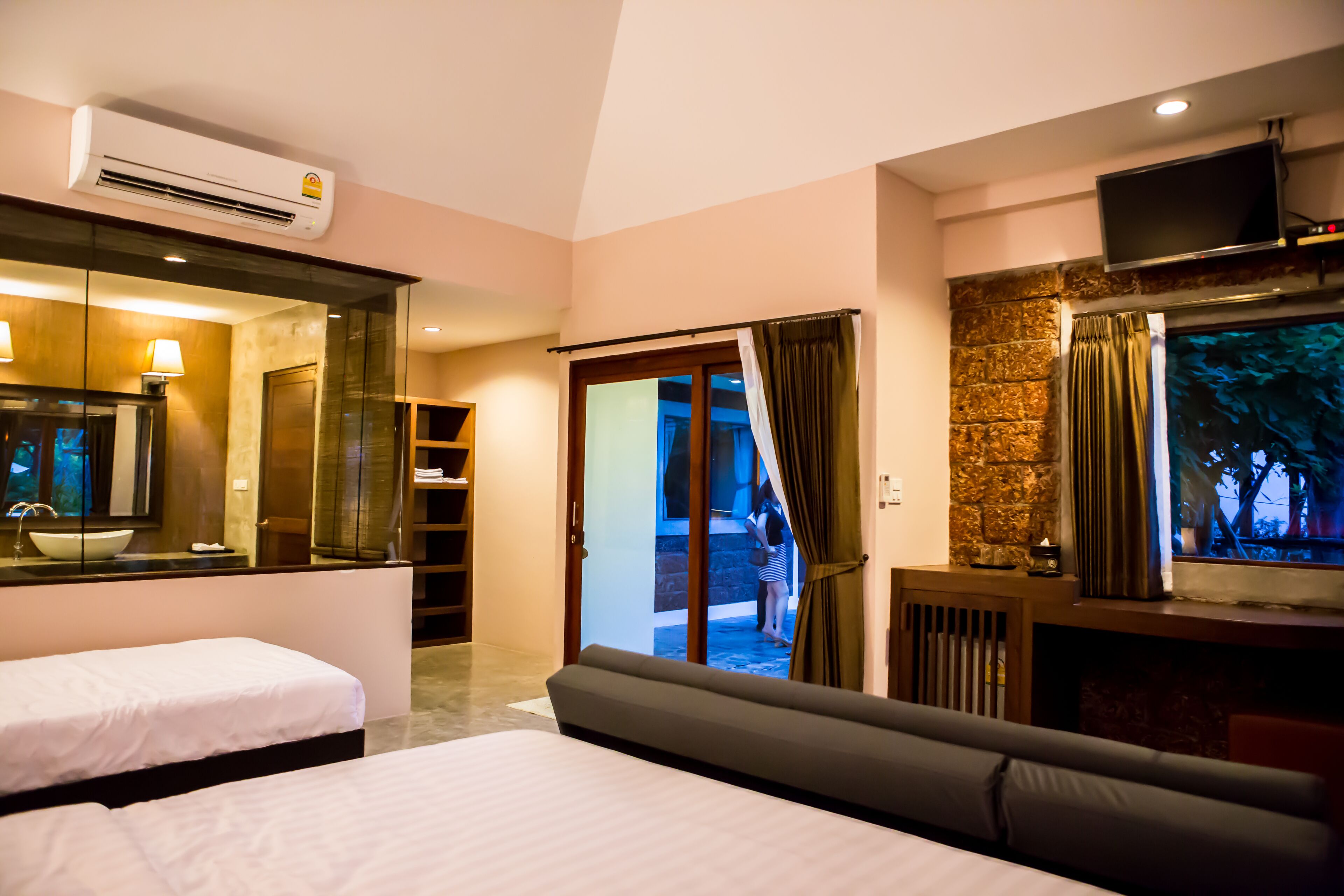 A hotel room with two beds, a TV, an air conditioner, a large mirror, and a view of the outside.