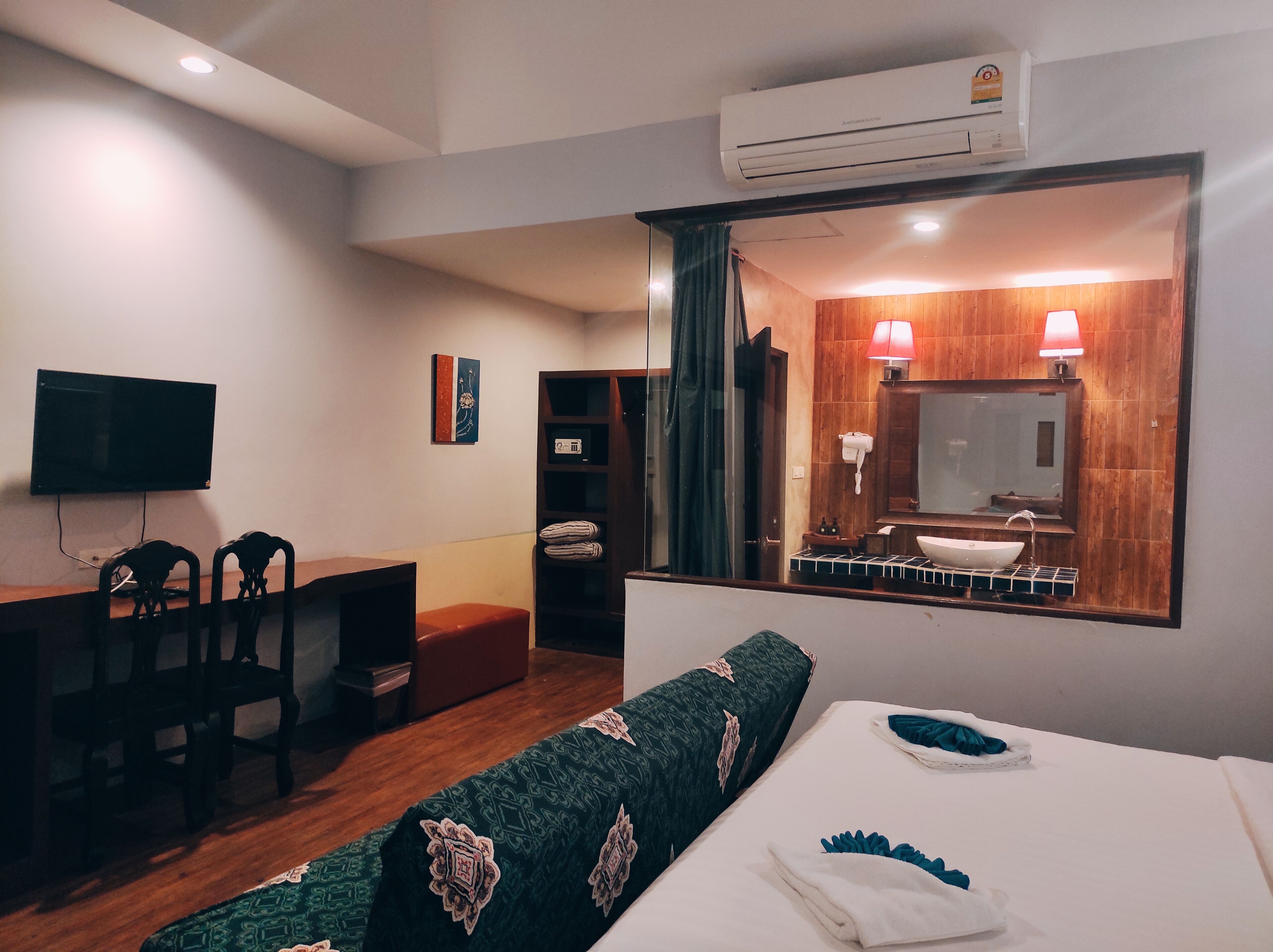 A hotel room with a bed, a desk, a chair, a television, a mirror, and an air conditioning unit.