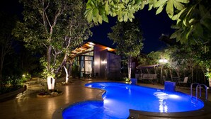 A swimming pool with a wooden deck and surrounding greenery, a modern building with glass walls, and outdoor seating.