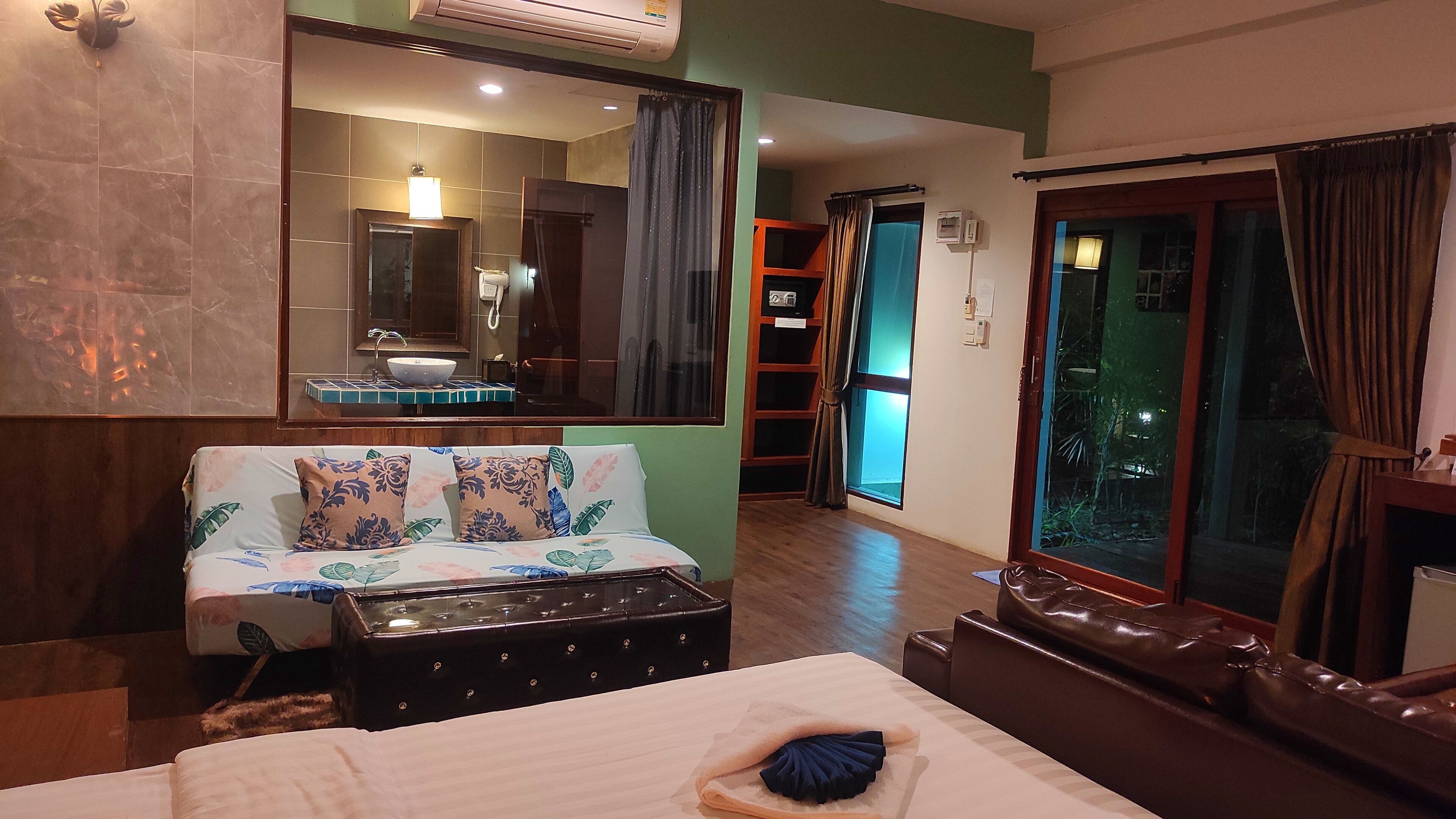 A hotel room with a bed, a sofa, a small table, a mirror, a window with curtains, and a bathroom with a sink and mirror.