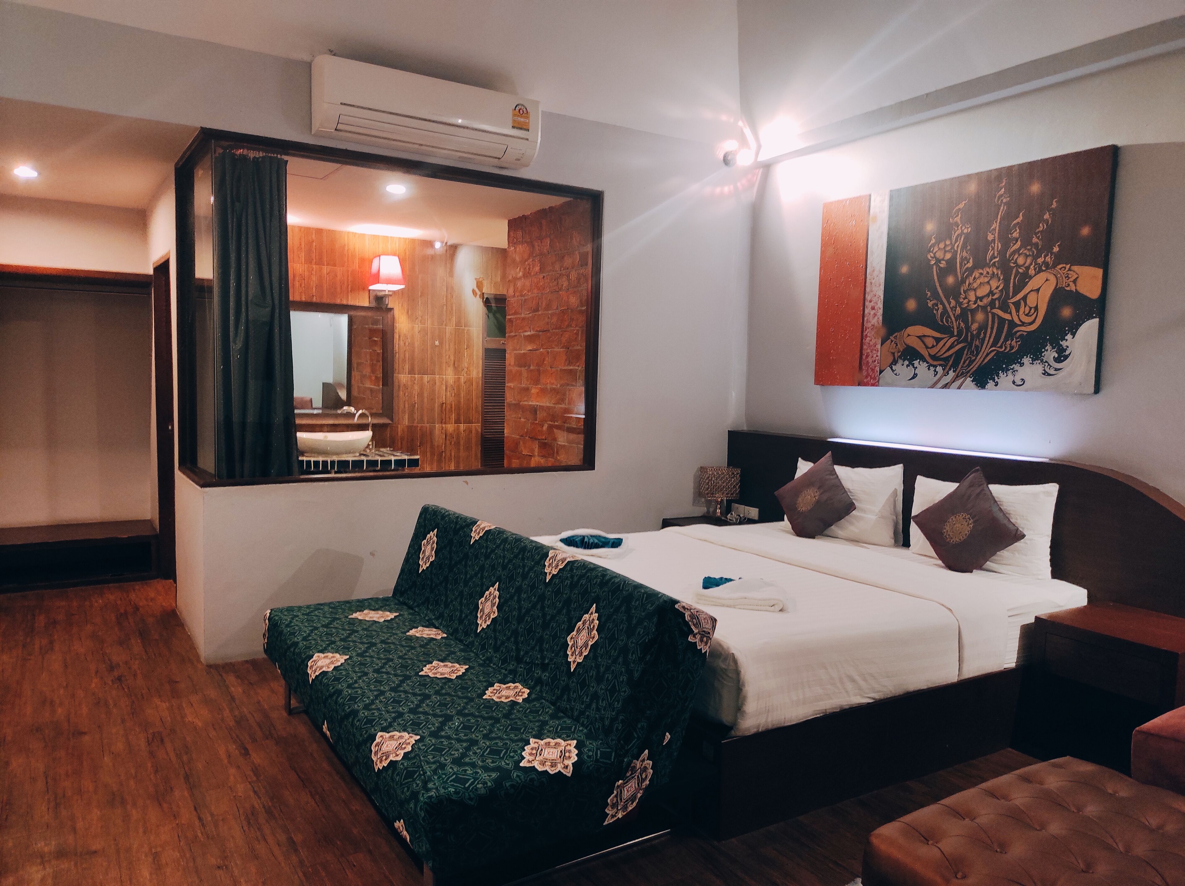 A hotel room with a bed, a bedside table, a mirror, an air conditioner, and a painting on the wall.